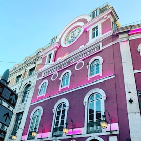 A tall building with a richly ornate exterior, painted in vibrant pink with white trim. The building has an inscription 'Teatro da Trindade' and features multiple arched windows with decorative moldings. Street lamps are visible, and a clear blue sky serves as the backdrop. A signpost with directional arrows and icons is also present in the image.