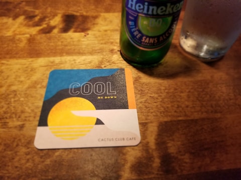 A wooden table holds a Heineken bottle and a glass filled with liquid. A square coaster from Cactus Club Cafe displays a design with a yellow sun, stylized waves, and a hand, carrying the text 'COOL ME DOWN' above it.