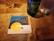 A wooden table holds a Heineken bottle and a glass filled with liquid. A square coaster from Cactus Club Cafe displays a design with a yellow sun, stylized waves, and a hand, carrying the text 'COOL ME DOWN' above it.