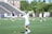 A female lacrosse player, wearing a white and light blue uniform, extends her arm with a lacrosse stick on a grassy field. In the background, other players and a coach are visible, along with a partially blurred grandstand and trees.