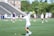 A female lacrosse player, wearing a white and light blue uniform, extends her arm with a lacrosse stick on a grassy field. In the background, other players and a coach are visible, along with a partially blurred grandstand and trees.