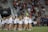 A large group of people, mainly young girls, are gathered on a sports field. Most are wearing matching outfits consisting of white shirts and dark shorts. The event appears to be in a stadium setting with many spectators seated in the background, indicating it may be a sporting or celebratory event.
