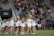 A large group of people, mainly young girls, are gathered on a sports field. Most are wearing matching outfits consisting of white shirts and dark shorts. The event appears to be in a stadium setting with many spectators seated in the background, indicating it may be a sporting or celebratory event.