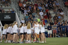 A large group of people, mainly young girls, are gathered on a sports field. Most are wearing matching outfits consisting of white shirts and dark shorts. The event appears to be in a stadium setting with many spectators seated in the background, indicating it may be a sporting or celebratory event.