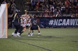 A group of female lacrosse players are intensely competing near the goal. One player is in possession of the ball, while two opponents are actively defending. The scene takes place on a well-lit field, surrounded by a crowd of spectators sitting on bleachers.