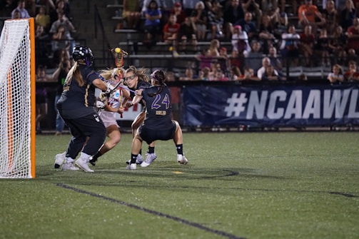 A group of female lacrosse players are intensely competing near the goal. One player is in possession of the ball, while two opponents are actively defending. The scene takes place on a well-lit field, surrounded by a crowd of spectators sitting on bleachers.