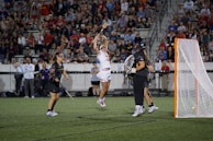 A lacrosse player in a white uniform leaps into the air to catch or throw a ball, surrounded by two players in dark uniforms. The scene takes place on a field with a crowd of spectators watching from the stands. A lacrosse goal is visible to the right.