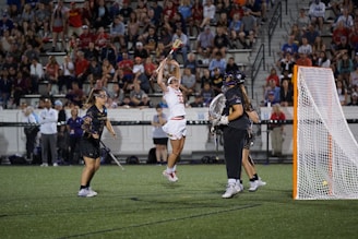 A lacrosse player in a white uniform leaps into the air to catch or throw a ball, surrounded by two players in dark uniforms. The scene takes place on a field with a crowd of spectators watching from the stands. A lacrosse goal is visible to the right.