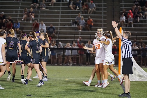 Players wearing sports uniforms are gathered on a lacrosse field. A team in dark uniforms and a team in white uniforms with one player wearing number 28 are seen celebrating near a goalpost. A referee in a striped shirt is raising an arm, indicating a call or score. Spectators are visible in the stands behind.
