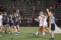 Players wearing sports uniforms are gathered on a lacrosse field. A team in dark uniforms and a team in white uniforms with one player wearing number 28 are seen celebrating near a goalpost. A referee in a striped shirt is raising an arm, indicating a call or score. Spectators are visible in the stands behind.
