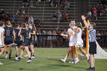 Players wearing sports uniforms are gathered on a lacrosse field. A team in dark uniforms and a team in white uniforms with one player wearing number 28 are seen celebrating near a goalpost. A referee in a striped shirt is raising an arm, indicating a call or score. Spectators are visible in the stands behind.
