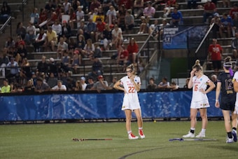 Athletes wearing white sports uniforms stand on a field with lacrosse sticks on the ground. They appear to be part of a lacrosse game, and a large audience is visible in the background seated on bleachers. The environment suggests a competitive sports event.