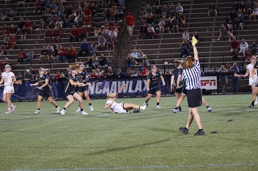A group of female athletes are engaged in a competitive lacrosse game on a well-lit field. One player in white is falling to the ground while holding a lacrosse stick. The opposing team in dark uniforms is surrounding her. A referee wearing a black and white striped shirt is raising an arm, possibly indicating a foul or penalty. In the background, a stadium filled with spectators watches the game, with the NCAA and ESPN logos visible.
