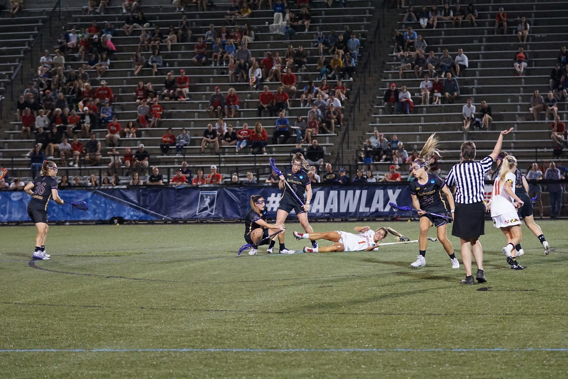 Athletes engaged in a women's lacrosse match, with several players in action as one falls to the ground. A referee is present on the field, and a crowd watches from the stadium benches in the background. The players are wearing uniforms, and the atmosphere appears competitive.