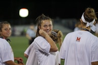 A group of young people wearing white shirts, possibly athletes, standing on a sports field under artificial lighting. One individual in the center is smiling directly at the camera, creating a friendly and engaging atmosphere.