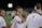 A group of young people wearing white shirts, possibly athletes, standing on a sports field under artificial lighting. One individual in the center is smiling directly at the camera, creating a friendly and engaging atmosphere.