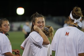 A group of young people wearing white shirts, possibly athletes, standing on a sports field under artificial lighting. One individual in the center is smiling directly at the camera, creating a friendly and engaging atmosphere.