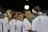 A group of young women wearing white sports jerseys with red and black accents are gathered together. One of them is smiling and wearing protective sports gear. They appear to be engaged in a conversation during nighttime on a sports field, as indicated by the blurred stadium lights in the background.