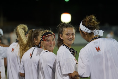 A group of young women wearing white sports jerseys with red and black accents are gathered together. One of them is smiling and wearing protective sports gear. They appear to be engaged in a conversation during nighttime on a sports field, as indicated by the blurred stadium lights in the background.