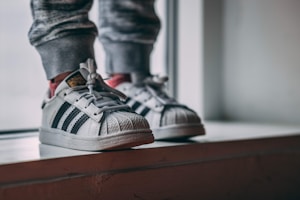 White sneakers with black stripes worn by a child, standing on a window ledge indoors. The legs are dressed in gray sweatpants with a slightly blurred background, suggesting a casual and cozy setting.