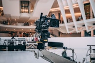 A professional video camera is mounted on a mechanical arm within a modern architectural setting. The background features sleek, curved structures with visible balconies and people observing the scene.