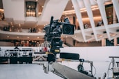 A professional video camera is mounted on a mechanical arm within a modern architectural setting. The background features sleek, curved structures with visible balconies and people observing the scene.