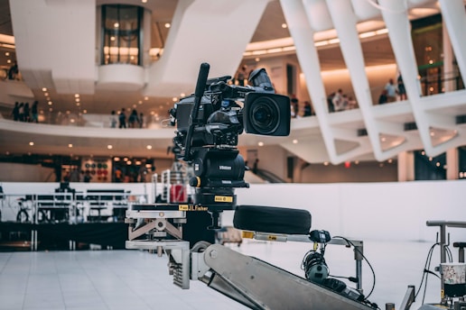 A professional video camera is mounted on a mechanical arm within a modern architectural setting. The background features sleek, curved structures with visible balconies and people observing the scene.