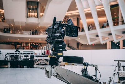 A professional video camera is mounted on a mechanical arm within a modern architectural setting. The background features sleek, curved structures with visible balconies and people observing the scene.
