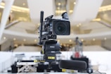 A professional broadcasting video camera mounted on a stable tripod within an indoor setting, possibly a modern auditorium or a large hall. The background is blurred, featuring an abstract architectural design with bright lights and a spacious interior.