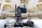 A professional broadcasting video camera mounted on a stable tripod within an indoor setting, possibly a modern auditorium or a large hall. The background is blurred, featuring an abstract architectural design with bright lights and a spacious interior.