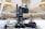 A professional broadcasting video camera mounted on a stable tripod within an indoor setting, possibly a modern auditorium or a large hall. The background is blurred, featuring an abstract architectural design with bright lights and a spacious interior.