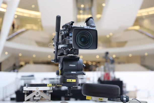 A professional broadcasting video camera mounted on a stable tripod within an indoor setting, possibly a modern auditorium or a large hall. The background is blurred, featuring an abstract architectural design with bright lights and a spacious interior.