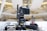 A professional broadcasting video camera mounted on a stable tripod within an indoor setting, possibly a modern auditorium or a large hall. The background is blurred, featuring an abstract architectural design with bright lights and a spacious interior.