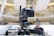 A professional broadcasting video camera mounted on a stable tripod within an indoor setting, possibly a modern auditorium or a large hall. The background is blurred, featuring an abstract architectural design with bright lights and a spacious interior.