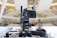 A professional broadcasting video camera mounted on a stable tripod within an indoor setting, possibly a modern auditorium or a large hall. The background is blurred, featuring an abstract architectural design with bright lights and a spacious interior.