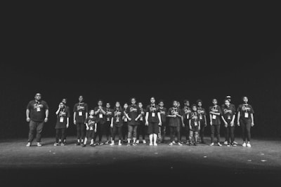 A group of people, mostly children, standing in a row on a stage. They are wearing matching black t-shirts with the word "Dare" printed on them and have badges hanging around their necks. The stage is dimly lit with a dark background, giving a sense of focus on the group.