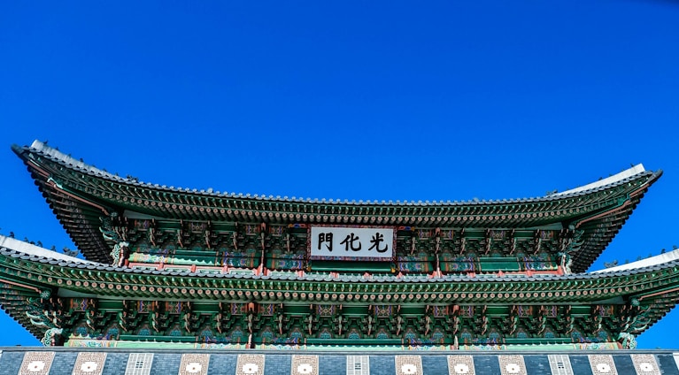 A traditional Korean architectural structure with intricate green and red painted details is set against a vivid blue sky. The roof features layered eaves with ornate patterns, and a sign with Korean characters is prominently displayed.