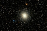 A dense cluster of stars forms a vibrant and luminous mass at the center of a dark backdrop. Numerous smaller stars are scattered throughout the surrounding space, with a mix of blue, red, and white hues.