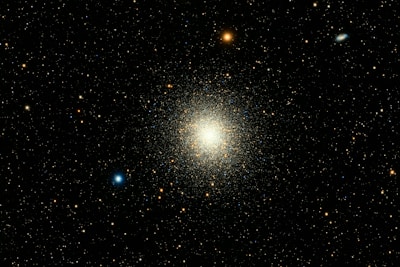 A dense cluster of stars forms a vibrant and luminous mass at the center of a dark backdrop. Numerous smaller stars are scattered throughout the surrounding space, with a mix of blue, red, and white hues.