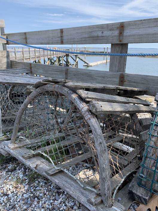 Weathered wooden lobster traps are stacked on top of a pebble-strewn surface beside a wooden dock. The ropes and netting are visible, with the calm blue water and a wooden pier in the background under a clear sky.