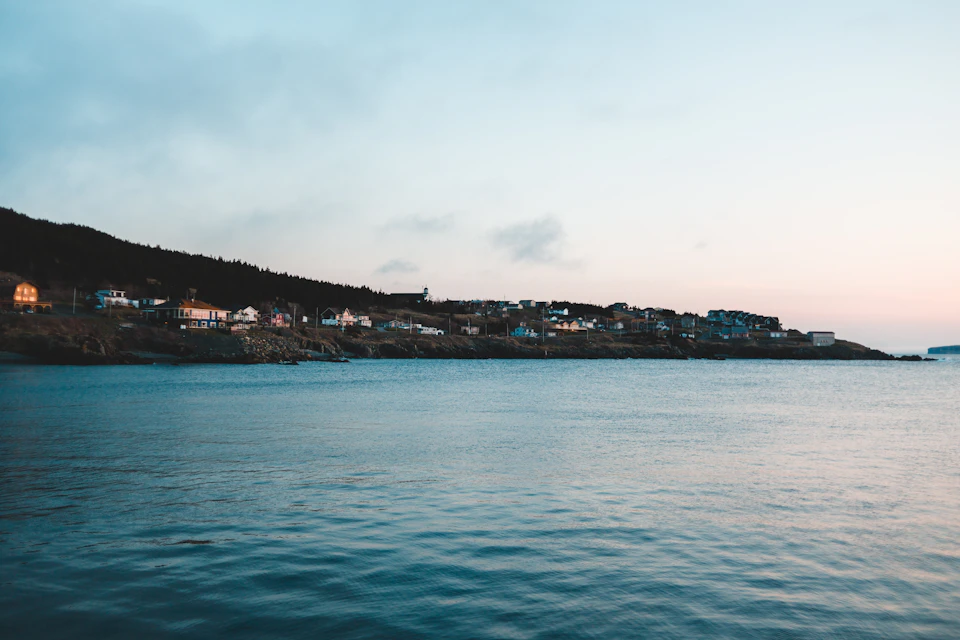 The image depicts a serene coastal town nestled on a hillside, with houses and buildings scattered across the landscape. The town is situated next to a body of water that stretches out towards the horizon, where it meets the sky at the edge of the frame. The colors in the scene are predominantly blue and green, reflecting the natural beauty of the environment. The perspective of the image is from the shoreline, looking out over the water, giving a sense of depth and vastness to the landscape.