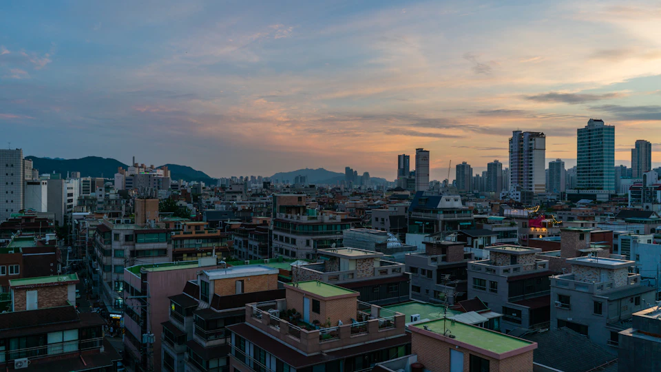 The image presents a panoramic view of a densely populated cityscape, with numerous buildings of varying heights and designs stretching out into the distance. The sky above is painted in hues of orange and pink, suggesting either sunrise or sunset, casting a warm glow over the urban landscape. In the foreground, there are several tall buildings that add to the density of the scene, while the city continues its expansion towards the horizon.