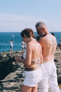 Two individuals in swimsuits stand near a rocky coastline with the sea in the background. Both have visible tattoos on their backs, and one person is holding a phone. Other people are seen in the distance near the water.