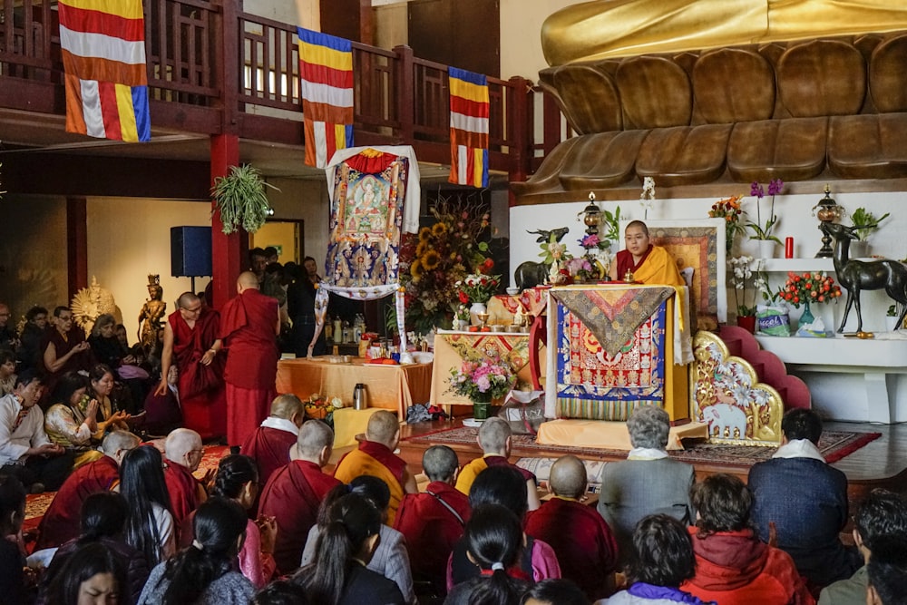 Dalai Lama sitting near table surrounded with people watching