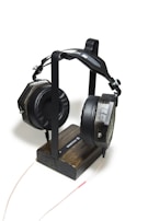 A pair of large, black headphones with cushioned ear pads is resting on a minimalist wooden headphone stand. The stand is constructed from dark-stained wood and has a metal frame that holds the headphones in place. A thin audio cable is connected to the headphones and extends towards the bottom of the image.