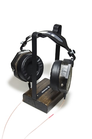 A pair of large, black headphones with cushioned ear pads is resting on a minimalist wooden headphone stand. The stand is constructed from dark-stained wood and has a metal frame that holds the headphones in place. A thin audio cable is connected to the headphones and extends towards the bottom of the image.