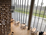 A modern library with a tall wall of books arranged in a grid pattern. Large floor-to-ceiling windows provide a view of an outdoor area with a pond and green grass. Minimalist wooden tables and chairs are scattered across the wooden floor, and a few people are seen sitting and reading.
