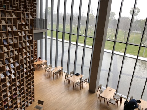 A modern library with a tall wall of books arranged in a grid pattern. Large floor-to-ceiling windows provide a view of an outdoor area with a pond and green grass. Minimalist wooden tables and chairs are scattered across the wooden floor, and a few people are seen sitting and reading.