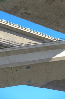 Two large concrete bridges intersecting at different angles against a clear blue sky. The structures feature sharp lines and strong geometric shapes with shadows cast at the intersection points. The concrete surfaces appear smooth and industrial.
