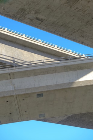 Two large concrete bridges intersecting at different angles against a clear blue sky. The structures feature sharp lines and strong geometric shapes with shadows cast at the intersection points. The concrete surfaces appear smooth and industrial.